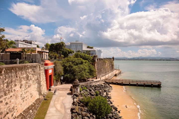 Coastal fort with stone walls, tropical greenery, and a wooden pier under a cloudy sky.