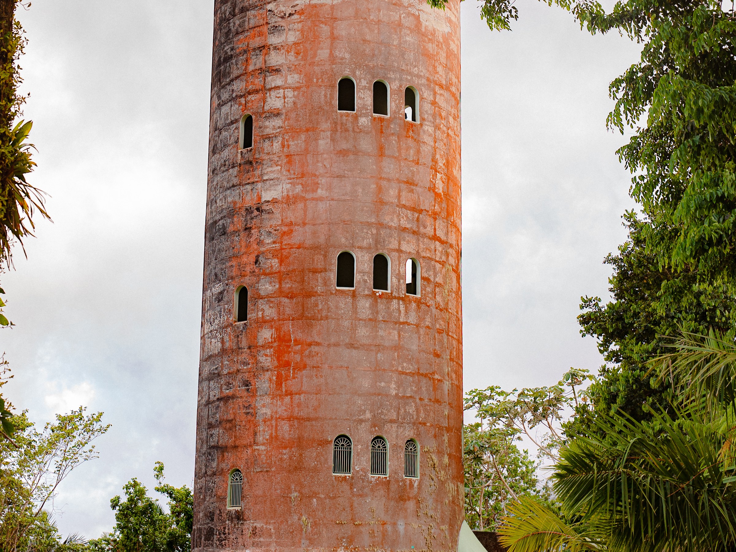 Historic round tower with small windows, surrounded by lush green trees on an overcast day.