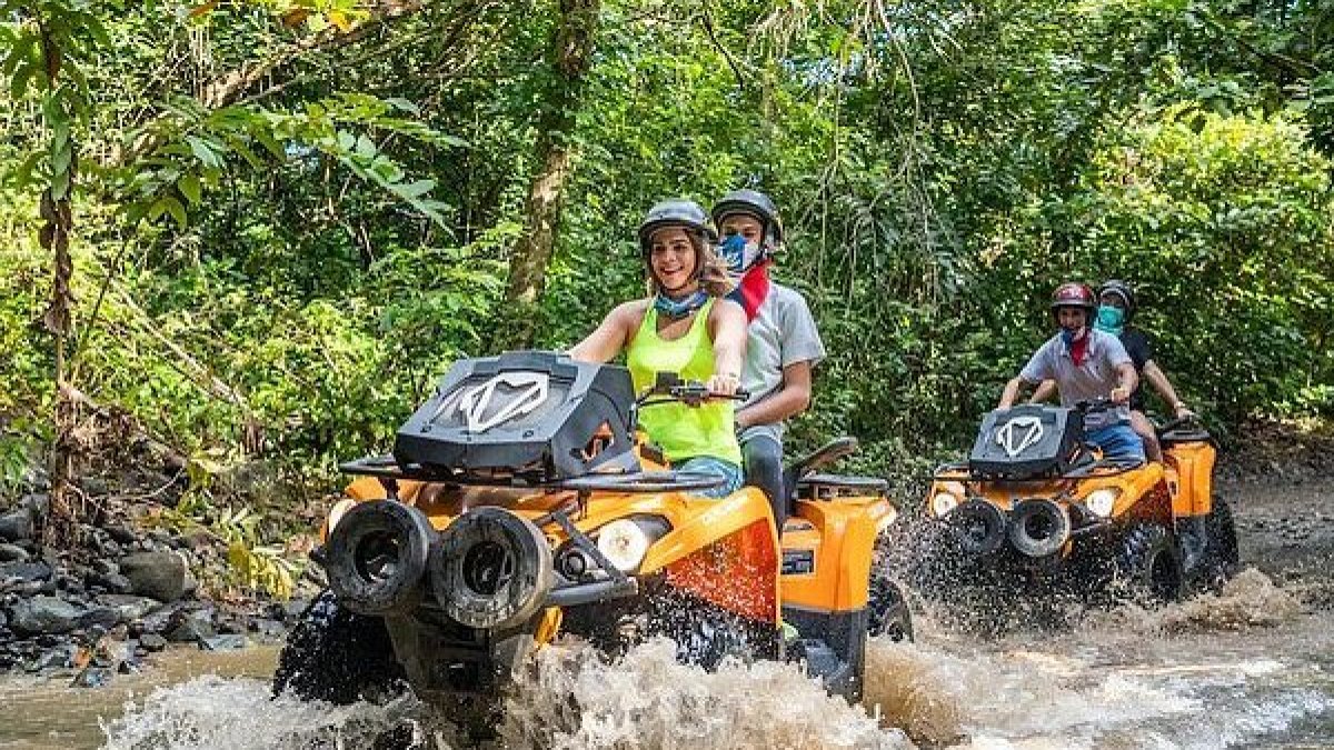 People riding orange ATVs through a muddy forest trail, splashing water.
