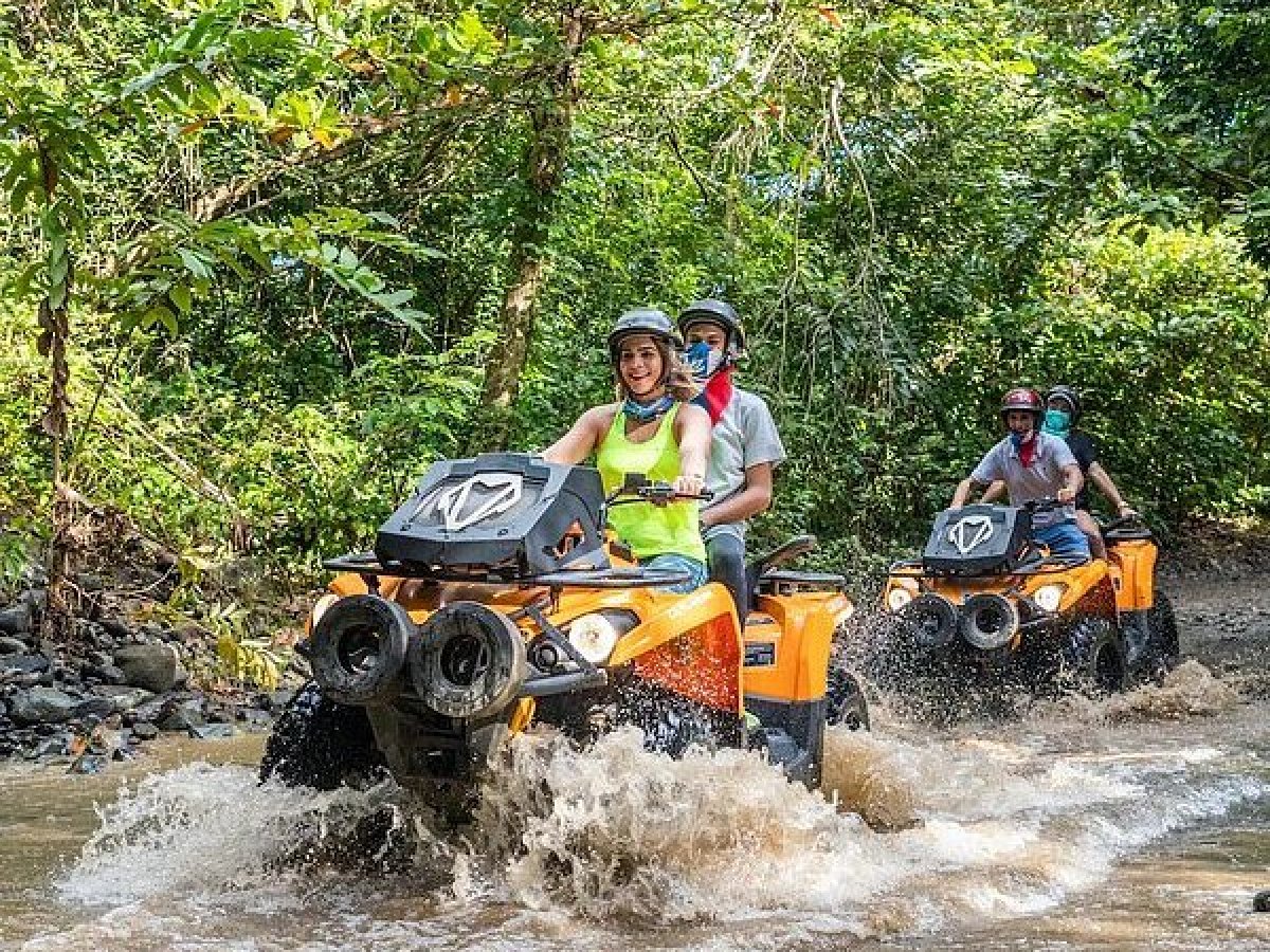 People riding orange ATVs through a muddy forest trail, splashing water.