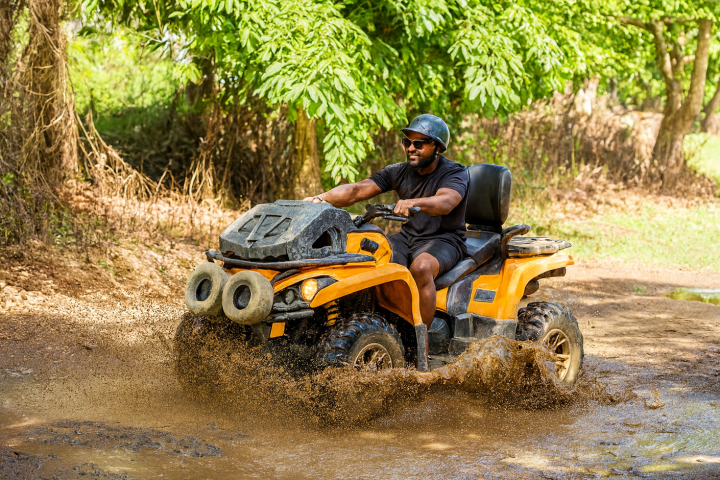 Man rides an orange quad bike through a muddy puddle in a forested area.