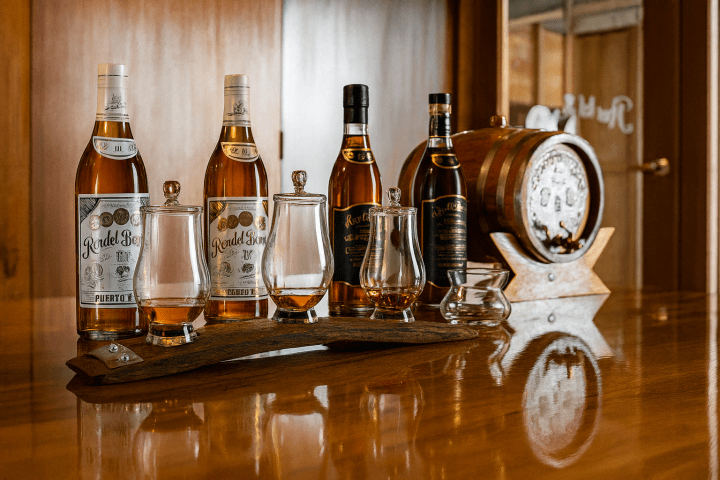 Whiskey bottles and glasses on a wooden table with a small barrel display in the background.