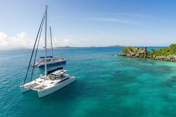 Two catamarans anchored near a rocky island in clear blue water under a blue sky.