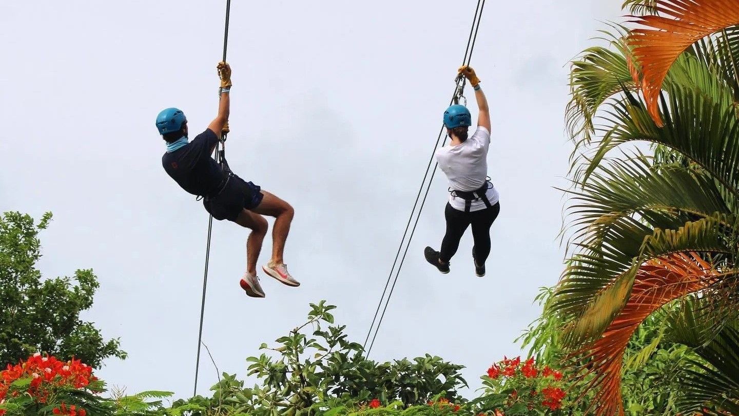 Two people zip-lining above lush greenery and orange flowers, wearing helmets and harnesses.