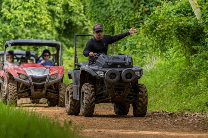 Person on ATV leading two off-road vehicles on a dirt path through a lush green forest.